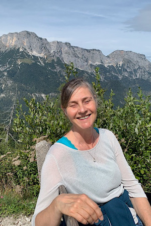 Frau sitzt auf Bank in der Sonne; lächelt mit geschlossenen Augen in die Kamera; Berge im Hintergrund